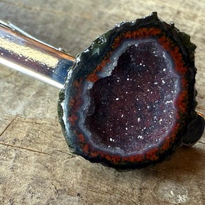 May include: A silver-toned metal tie clip featuring a circular geode slice. The geode has a dark, rough outer edge, a vibrant orange-red inner ring, and a deep purple, crystalline center with small, sparkling white mineral deposits.