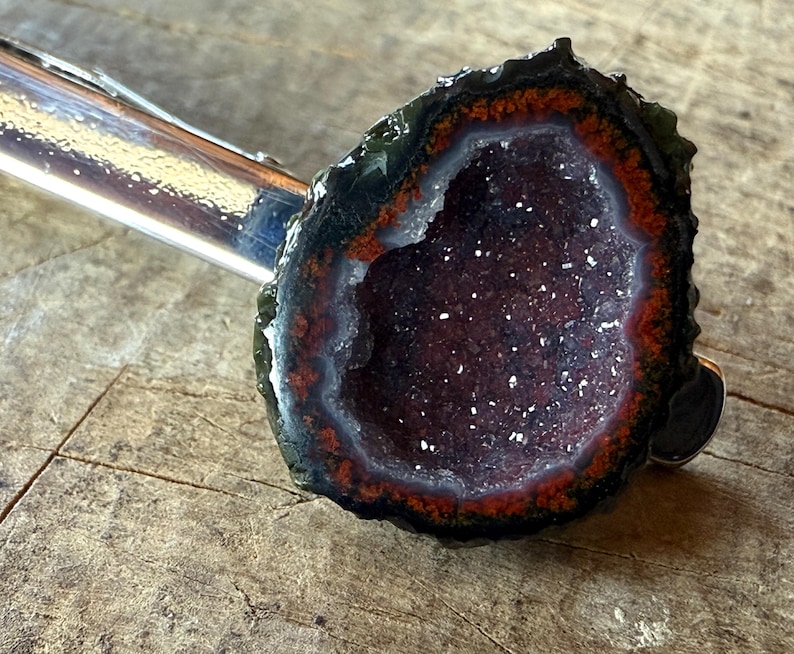 May include: A silver-toned metal tie clip featuring a circular geode slice. The geode has a dark, rough outer edge, a vibrant orange-red inner ring, and a deep purple, crystalline center with small, sparkling white mineral deposits.
