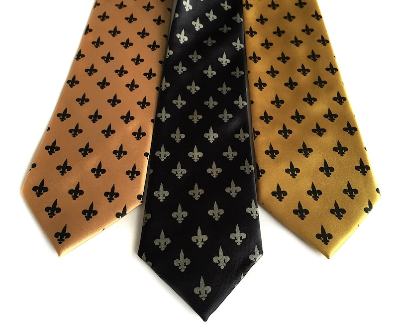 May include: Three silk ties with a fleur-de-lis pattern. One tie is black with silver fleur-de-lis, one is gold with black fleur-de-lis, and one is gold with black fleur-de-lis.
