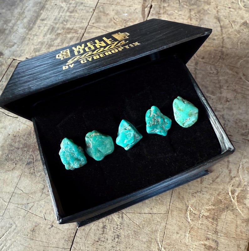 May include: A black wooden box with gold lettering reading "WELL DONE" and "BY CYBEROPTIX" contains five turquoise gemstones. The gemstones are a vibrant turquoise color and have a rough, natural texture. The box is open, revealing the gemstones.