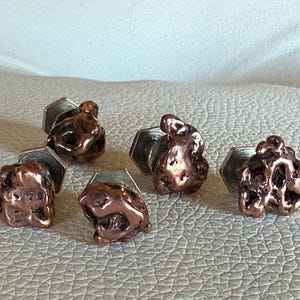 May include: Six copper-colored cufflinks with a textured, nugget-like design. Each cufflink is attached to a silver-colored, hexagonal base. The cufflinks are arranged on a light-colored, textured surface.
