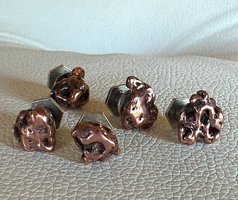 May include: Six copper-colored cufflinks with a textured, nugget-like design. Each cufflink is attached to a silver-colored, hexagonal base. The cufflinks are arranged on a light-colored, textured surface.