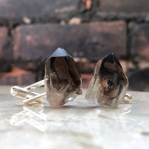 May include: A pair of silver cufflinks with smoky quartz crystal accents. The crystals are faceted and have a dark gray color.