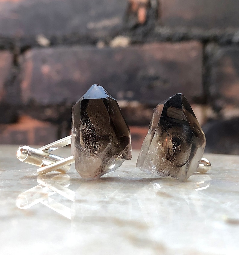 May include: A pair of silver cufflinks with smoky quartz crystal accents. The crystals are faceted and have a dark gray color.