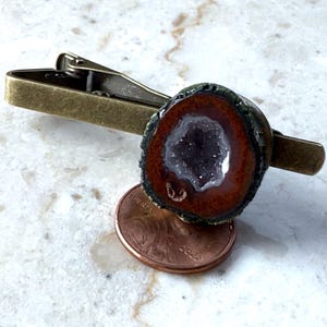 May include: A bronze-toned metal tie clip featuring a circular geode slice with a dark brown outer ring and a sparkling purple amethyst center, positioned directly above a copper penny for scale to show the size of the stone ornament.
