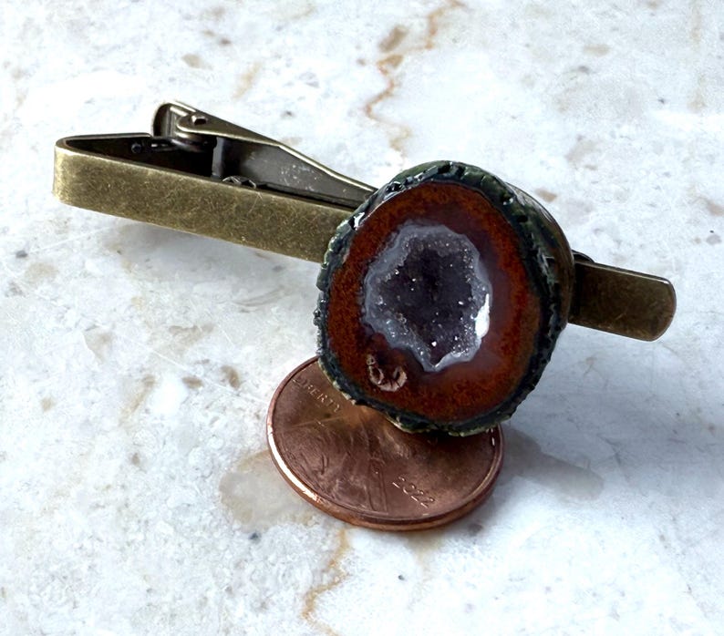 May include: A bronze-toned metal tie clip featuring a circular geode slice with a dark brown outer ring and a sparkling purple amethyst center, positioned directly above a copper penny for scale to show the size of the stone ornament.