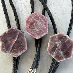 May include: A bolo tie featuring three hexagonal, raw, purple-toned mineral stones attached to black braided leather cords. The stones have a rough, crystalline texture with visible surface fissures and natural variations in their deep mauve color.