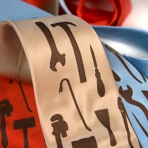 May include: Three neckties with different tool designs. The first tie is light blue with a wrench, hammer, and screwdriver design. The second tie is beige with a hammer, crowbar, screwdriver, and putty knife design. The third tie is red with a wrench, hammer, and screwdriver design.
