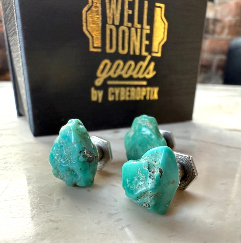 May include: Three turquoise stone cufflinks with silver-colored metal hardware. The cufflinks are displayed in front of a black box with gold lettering that reads "WELL DONE goods by CYBEROPTIX". The turquoise stones have a rough, natural texture.