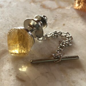 May include: A silver tie tack with a gold-toned, faceted, crystal-like stone. The tie tack has a chain and a bar for attaching to clothing.