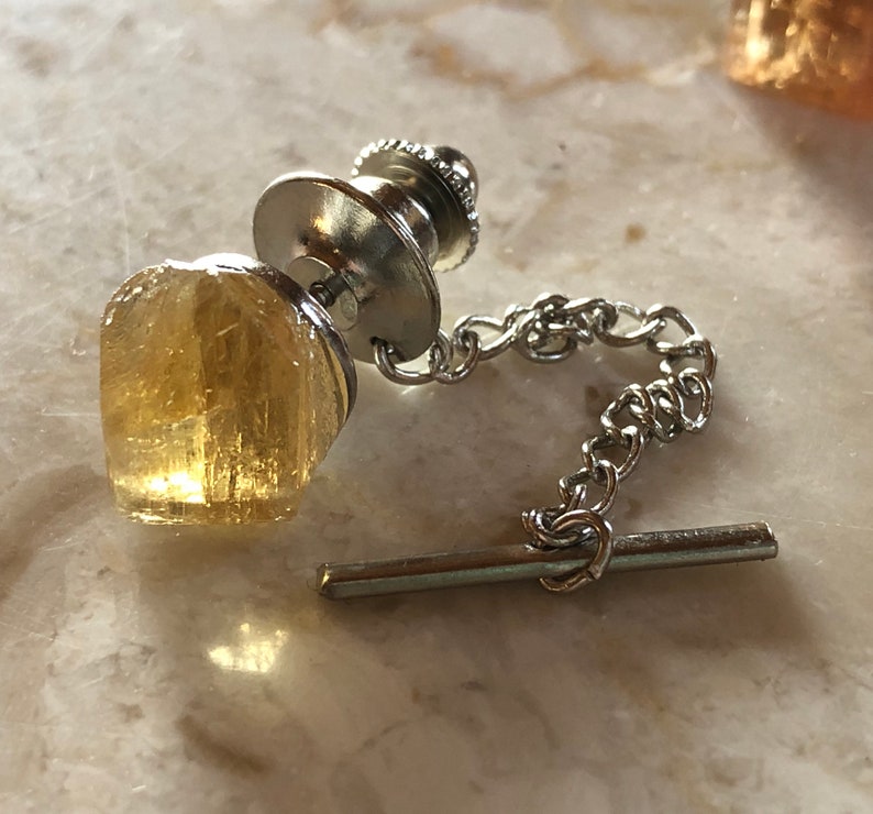 May include: A silver tie tack with a gold-toned, faceted, crystal-like stone. The tie tack has a chain and a bar for attaching to clothing.