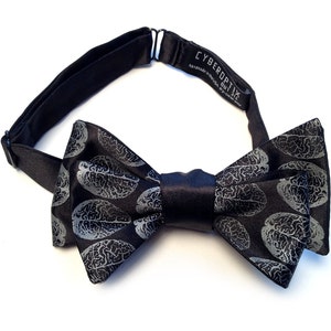 May include: A black bow tie with a silver brain pattern. The bow tie has a black adjustable strap with the text "CYBEROPTIX" printed on it.