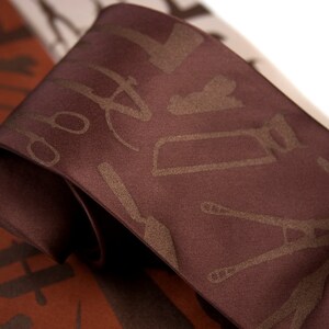 May include: A brown silk tie with a pattern of tools in a lighter brown color. The tools include a saw, a wrench, a hammer, and a screwdriver.