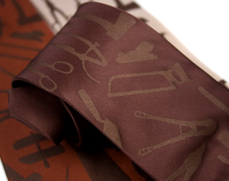 May include: A brown silk tie with a pattern of tools in a lighter brown color. The tools include a saw, a wrench, a hammer, and a screwdriver.