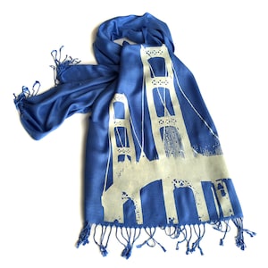 May include: A blue silk scarf with a white silhouette of a bridge printed on it. The scarf has fringe on the bottom edge.