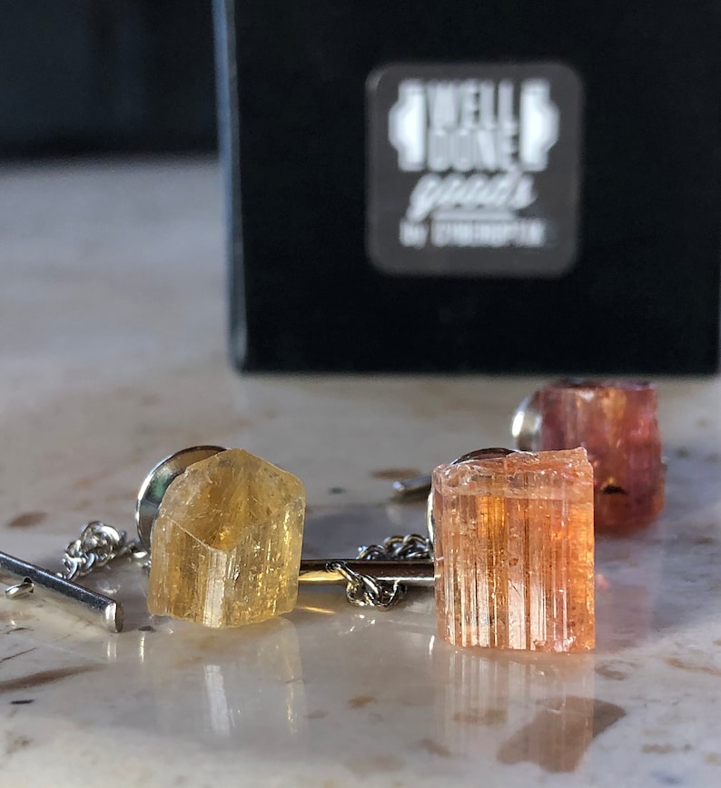 May include: Two gold and orange crystal tie tacks with silver chains. The tie tacks are set against a white background.
