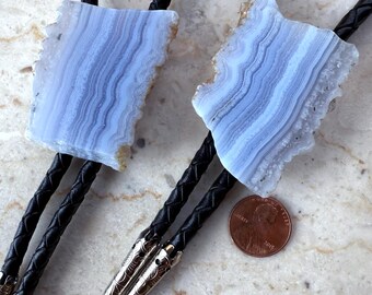 May include: Two blue lace agate bolo ties with black braided cords and silver-toned tips. The agate stones have a light blue and white banded pattern. A penny is included for size reference.