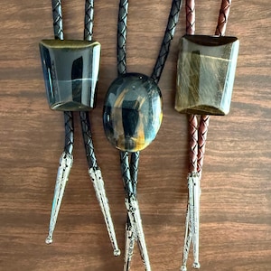 May include: Three bolo ties featuring polished gemstones. Two ties have trapezoidal stones in dark brown and gold, while the center tie has an oval blue and gold stone. Each tie includes a braided leather cord and silver-toned metal tips with engravings.