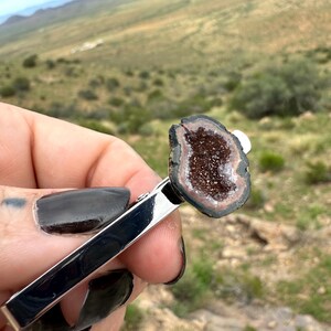 May include: A silver-toned metal tie clip featuring a polished, irregular-shaped geode slice at one end. The geode has a dark brown, crystalline center surrounded by concentric rings of black and tan mineral banding, creating a natural contrast.