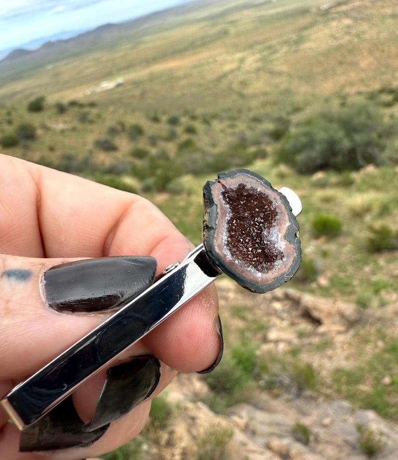May include: A silver-toned metal tie clip featuring a polished, irregular-shaped geode slice at one end. The geode has a dark brown, crystalline center surrounded by concentric rings of black and tan mineral banding, creating a natural contrast.