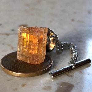 May include: A gold-toned tie tack with a clear, faceted, orange crystal. The crystal is attached to a silver chain and a bar.