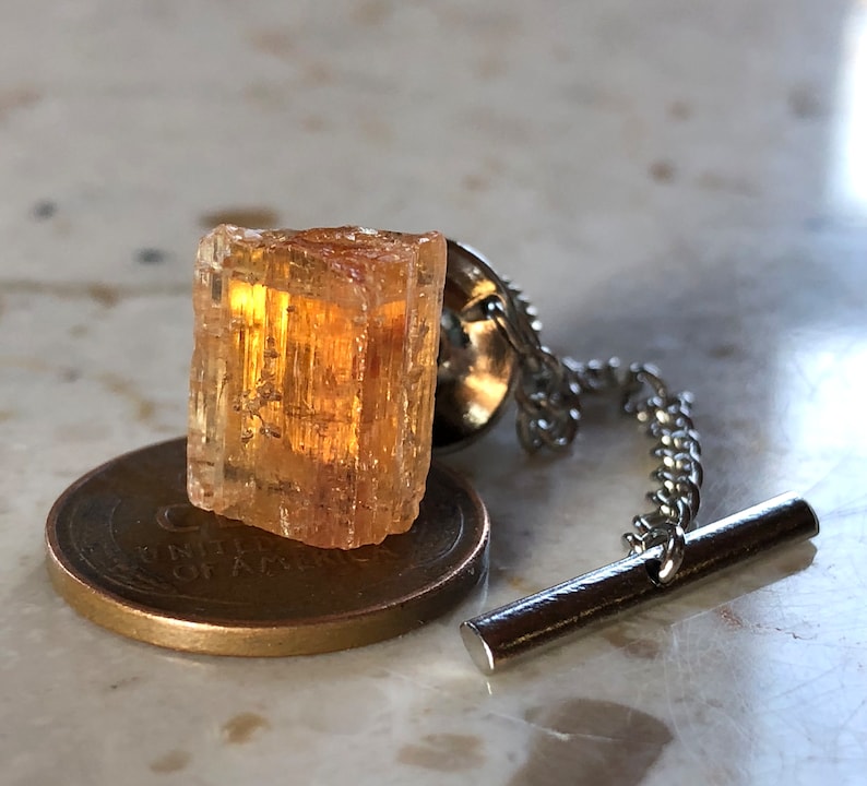 May include: A gold-toned tie tack with a clear, faceted, orange crystal. The crystal is attached to a silver chain and a bar.