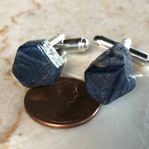 May include: A pair of cufflinks featuring raw blue sapphire gemstones. The sapphires are set in silver cufflink backs.