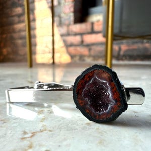 May include: A silver-toned metal tie clip featuring a circular geode slice at one end. The geode has a dark, black outer edge, a reddish-brown ring, and a deep, sparkling purple crystalline center, creating a distinct contrast against the polished metal.
