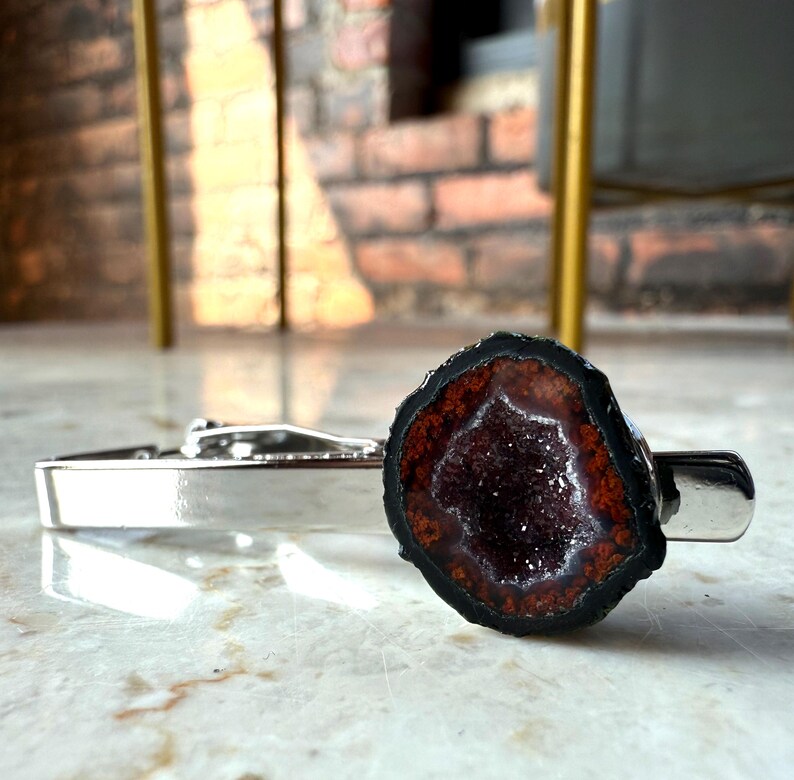 May include: A silver-toned metal tie clip featuring a circular geode slice at one end. The geode has a dark, black outer edge, a reddish-brown ring, and a deep, sparkling purple crystalline center, creating a distinct contrast against the polished metal.