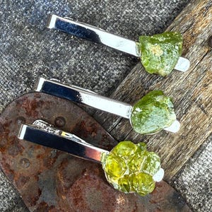 May include: Three silver tie clips, each adorned with a rough-cut, light green peridot gemstone. The clips are arranged on a textured, rustic surface, showcasing the natural beauty of the gemstones and the sleek design of the clips.