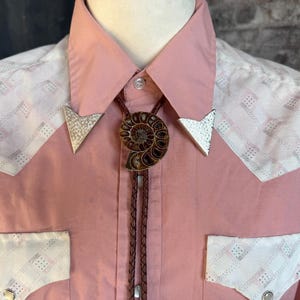 A pink Western-style shirt with white patterned accents on the shoulders and pockets. The shirt features a bolo tie with a brown ammonite fossil pendant and silver arrow-shaped tips. The shirt is buttoned up.