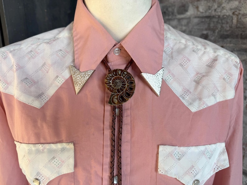 A pink Western-style shirt with white patterned accents on the shoulders and pockets. The shirt features a bolo tie with a brown ammonite fossil pendant and silver arrow-shaped tips. The shirt is buttoned up.
