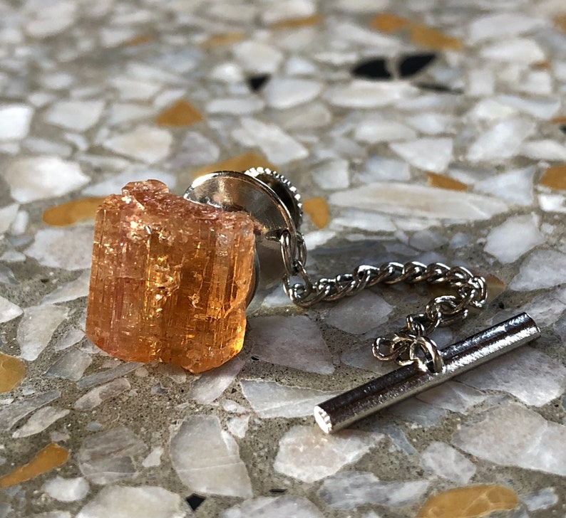 May include: A brown and orange crystal on a silver tie tack with a chain.