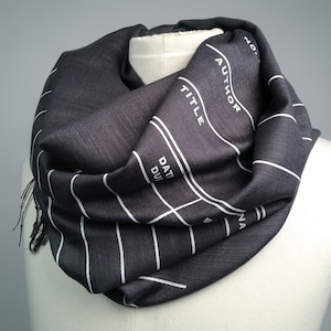 May include: A black and white striped infinity scarf with a film slate design. The scarf features the words "AUTHOR", "TITLE", "DATE DUE", and "SCENE".