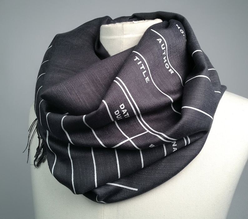 May include: A black and white striped infinity scarf with a film slate design. The scarf features the words "AUTHOR", "TITLE", "DATE DUE", and "SCENE".