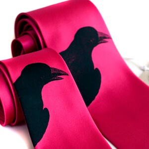 以下が含まれることがあります： 黒いカラスのシルエットがプリントされたピンクのシルクネクタイ2本。ネクタイは巻かれて一緒に結ばれています。