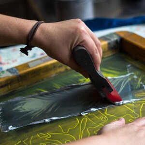 May include: A person using a red and black squeegee to apply silver ink to a screen printing frame. The frame has a yellow design on it.