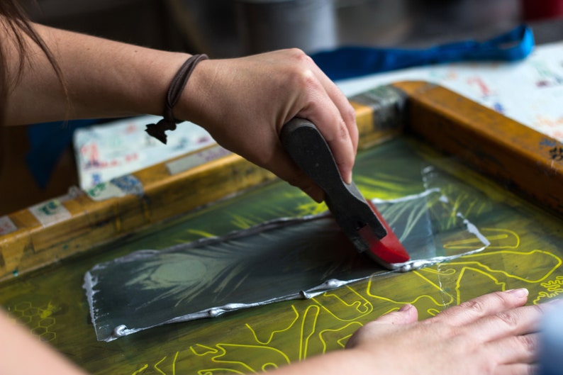 May include: A person using a red and black squeegee to apply silver ink to a screen printing frame. The frame has a yellow design on it.