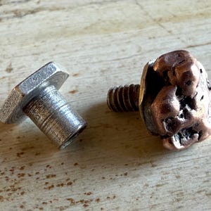 May include: Two metal hardware pieces. One is a silver-colored bolt with a hexagonal head. The other is a copper-colored bolt with a textured, nugget-like head. Both are on a light-colored wooden surface.