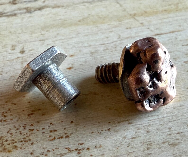 May include: Two metal hardware pieces. One is a silver-colored bolt with a hexagonal head. The other is a copper-colored bolt with a textured, nugget-like head. Both are on a light-colored wooden surface.