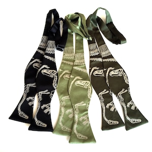May include: Three bow ties with a dinosaur skeleton print. One bow tie is black with a white print, and the other two are green with a white print.