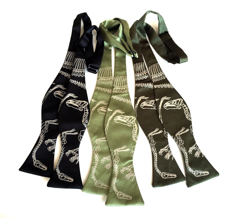 May include: Three bow ties with a dinosaur skeleton print. One bow tie is black with a white print, and the other two are green with a white print.