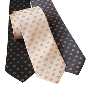 Puede incluir: Tres corbatas con diferentes patrones. Una corbata es de color beige claro con un patr&oacute;n repetido de peque&ntilde;os gatos naranjas. Las otras dos corbatas son de color gris oscuro con un patr&oacute;n repetido de peque&ntilde;os gatos grises.