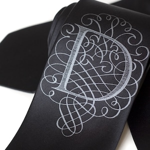 Può includere: Una cravatta di seta nera con un monogramma argentato della lettera "D" in un carattere di scrittura decorativo.