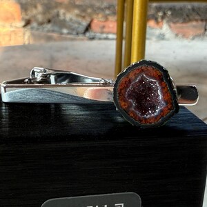 May include: A silver-toned metal tie clip featuring a circular geode slice at one end. The geode has a dark, crystalline center surrounded by a ring of deep orange-red mineral deposits, all encased within a thin, dark, natural outer edge.