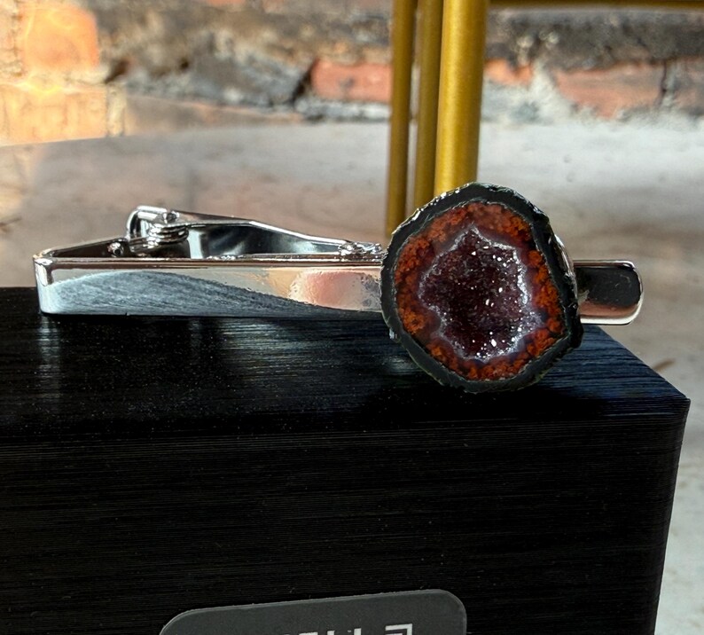 May include: A silver-toned metal tie clip featuring a circular geode slice at one end. The geode has a dark, crystalline center surrounded by a ring of deep orange-red mineral deposits, all encased within a thin, dark, natural outer edge.