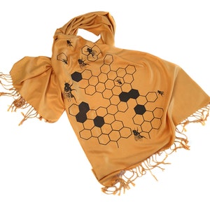 May include: A mustard yellow scarf with a black honeycomb pattern and bees. The scarf has fringe on the ends.