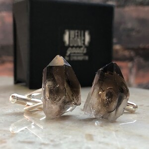 May include: A pair of cufflinks featuring two smoky quartz crystals, each with a unique shape and texture. The cufflinks are made of silver and have a polished finish.