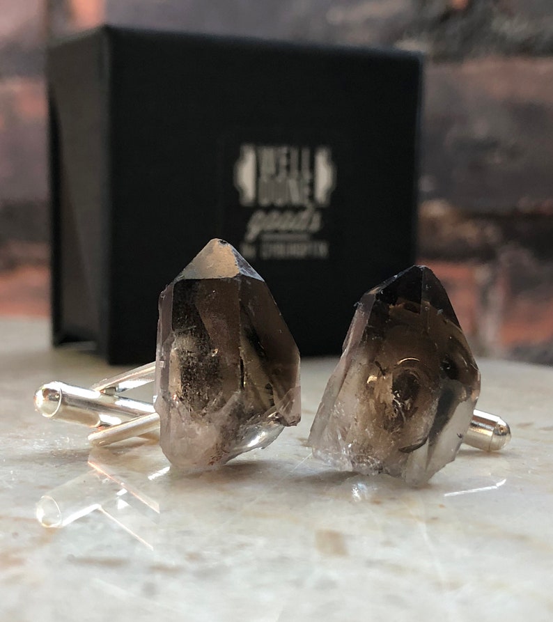 May include: A pair of cufflinks featuring two smoky quartz crystals, each with a unique shape and texture. The cufflinks are made of silver and have a polished finish.
