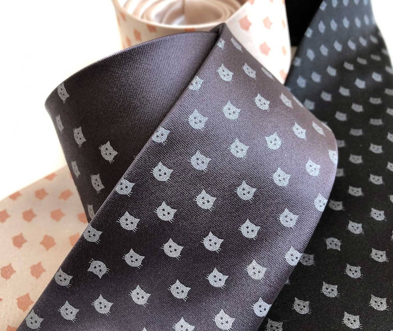 Puede incluir: Tres corbatas con un patr&oacute;n repetido de caras de gatos grises sobre un fondo gris oscuro. Una corbata es negra con un patr&oacute;n repetido de caras de gatos grises sobre un fondo negro.